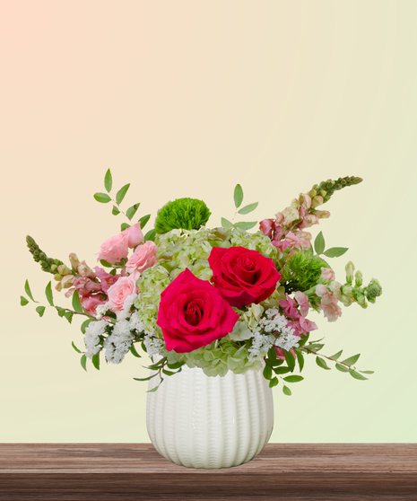 She’s All That! floral arrangement with hot pink roses, blush spray roses, green hydrangeas, snapdragons, and green dianthus in a white pedestal vase.