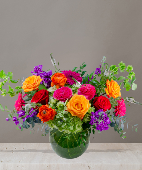Zara floral arrangement featuring jewel-toned roses, ranunculus, hydrangeas, and gerbera daisies in a round glass vase with tropical leaf lining.