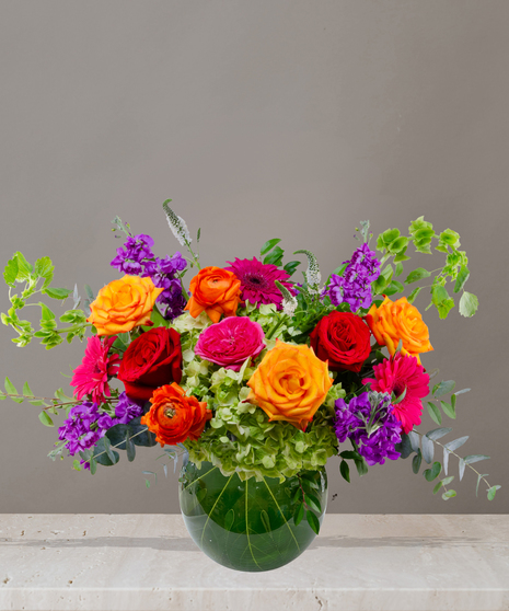 Zara floral arrangement featuring jewel-toned roses, ranunculus, hydrangeas, and gerbera daisies in a round glass vase with tropical leaf lining.