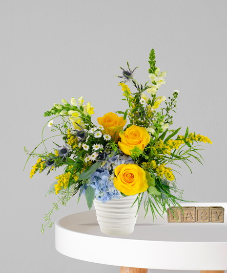 Pride & Boy flower arrangement with yellow roses, blue hydrangeas, and wildflowers in a white ceramic vase for a baby boy