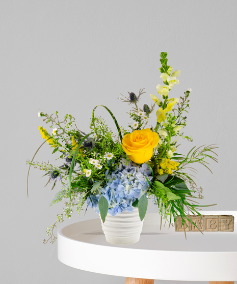 Pride & Boy flower arrangement with yellow roses, blue hydrangeas, and wildflowers in a white ceramic vase for a baby boy