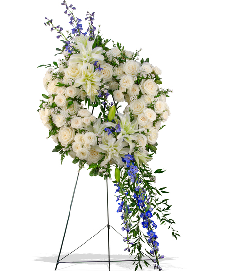 Elegant sympathy wreath with cascading white lilies, blue delphinium, ivory roses, and greenery on a metal easel – Touch of Color Collection by Billy Heroman’s