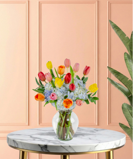 April Showers exclusive Easter flower arrangement with colorful red, pink, yellow, and orange tulips bursting from a base of blue hydrangeas, set in a clear glass flared vase