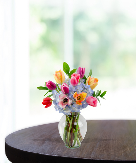 Tulip Treasures floral arrangement featuring a colorful variety of red, pink, yellow, and purple tulips mixed with blue hydrangeas in a clear glass vase.