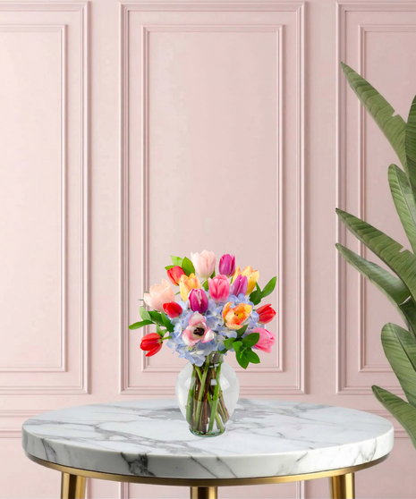 Burst of Joy Mother's Day flower arrangement featuring a colorful variety of red, pink, yellow, and purple tulips mixed with blue hydrangeas in a clear glass vase.