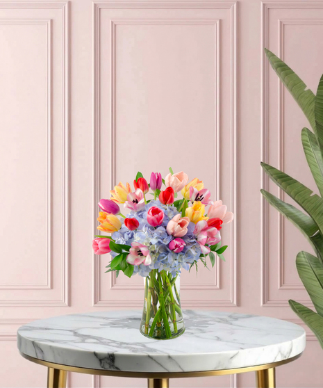 Burst of Joy Mother's Day flower arrangement featuring a colorful variety of red, pink, yellow, and purple tulips mixed with blue hydrangeas in a clear glass vase.