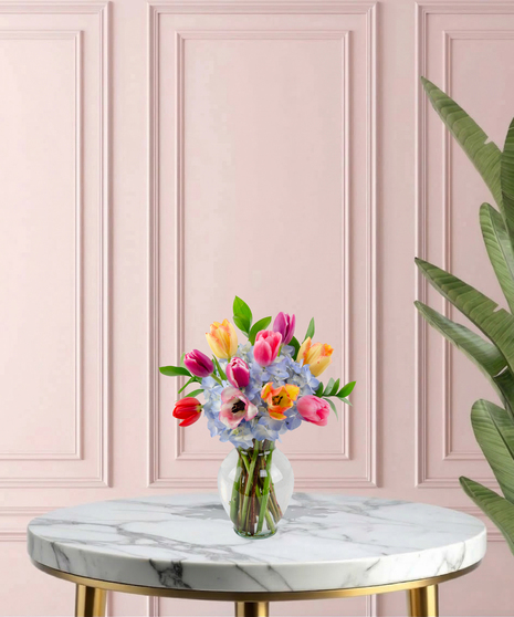 Burst of Joy Mother's Day flower arrangement featuring a colorful variety of red, pink, yellow, and purple tulips mixed with blue hydrangeas in a clear glass vase.