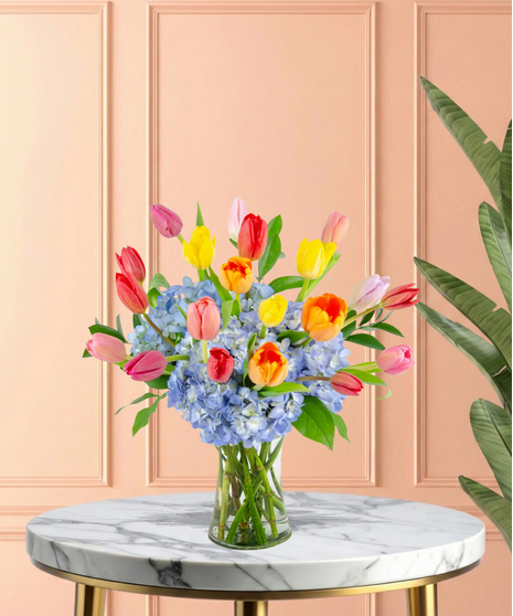 April Showers exclusive Easter flower arrangement with colorful red, pink, yellow, and orange tulips bursting from a base of blue hydrangeas, set in a clear glass flared vase