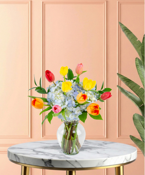 April Showers exclusive Easter flower arrangement with colorful red, pink, yellow, and orange tulips bursting from a base of blue hydrangeas, set in a clear glass flared vase