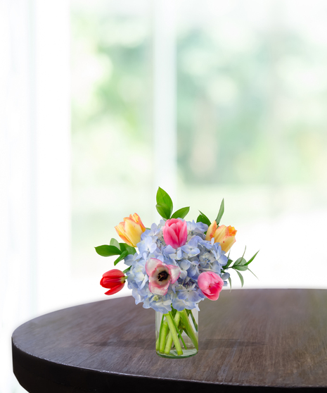 Tulip Treasures floral arrangement featuring a colorful variety of red, pink, yellow, and purple tulips mixed with blue hydrangeas in a clear glass vase.