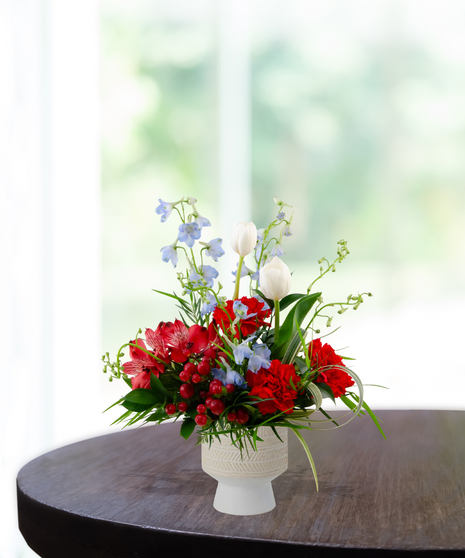 U.S.Yay patriotic flower arrangement featuring white tulips, blue delphinium, red carnations, and red berries in a textured white pedestal vase.