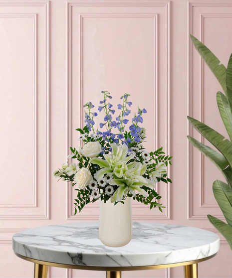 Under Her Wing Mother's Day flower arrangement featuring white roses, white lilies, blue delphinium, and blue hydrangeas in a matte white vase.