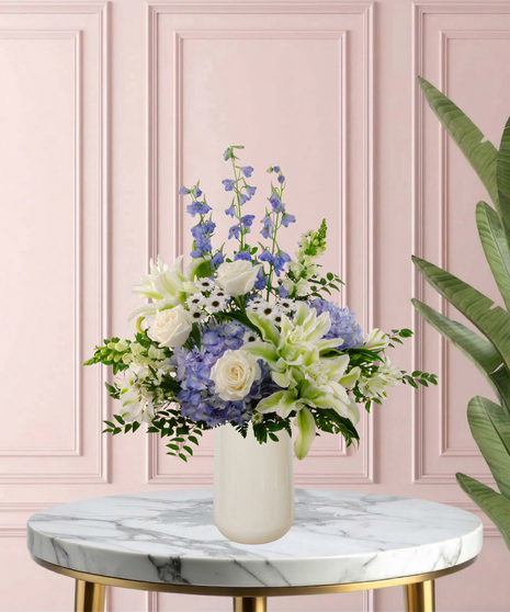 Under Her Wing Mother's Day flower arrangement featuring white roses, white lilies, blue delphinium, and blue hydrangeas in a matte white vase.