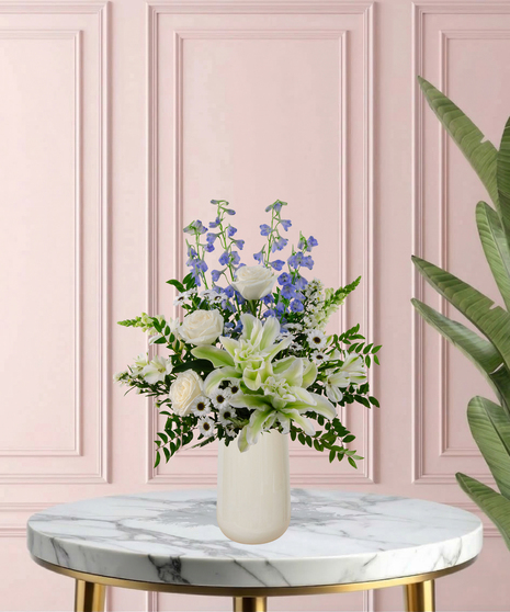 Under Her Wing Mother's Day flower arrangement featuring white roses, white lilies, blue delphinium, and blue hydrangeas in a matte white vase.