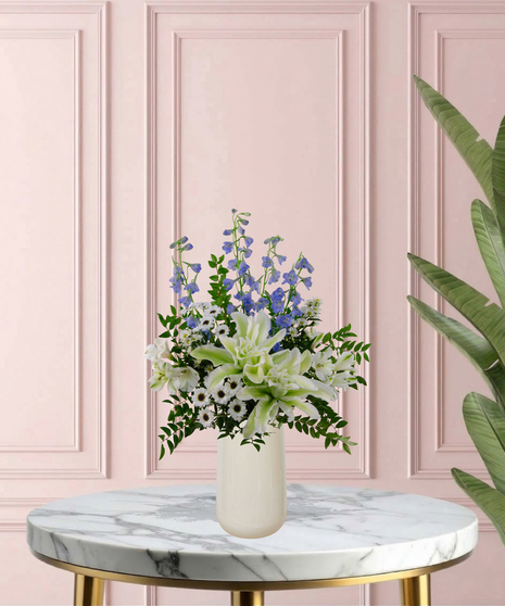 Under Her Wing Mother's Day flower arrangement featuring white roses, white lilies, blue delphinium, and blue hydrangeas in a matte white vase.