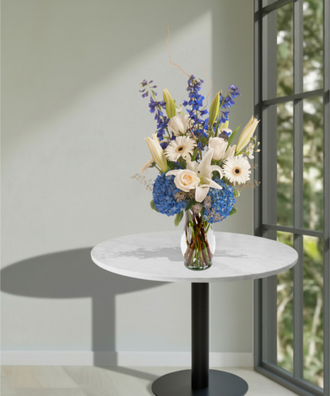 Sympathy vase with white roses, lilies, and blue hydrangeas, accented with blue ribbon and curly willow