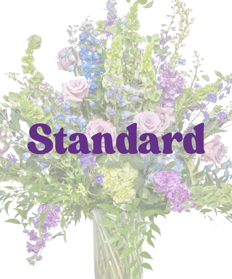 Pastel spring funeral flower arrangement in a tall glass vase with lavender roses, blue delphinium, green hydrangea, and bells of Ireland by Billy Heroman’s in Baton Rouge, LA
