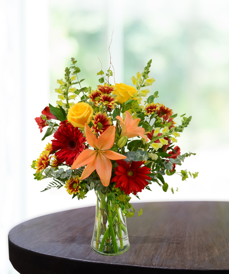 Bright fall flower arrangement with orange lilies, yellow roses, red gerberas, and seasonal greens in a clear vase