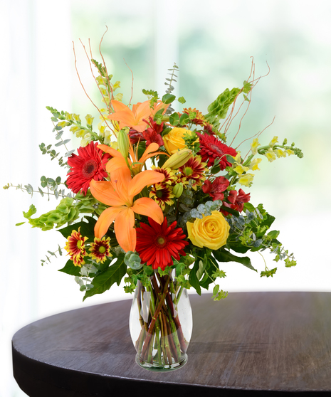 Bright fall flower arrangement with orange lilies, yellow roses, red gerberas, and seasonal greens in a clear vase