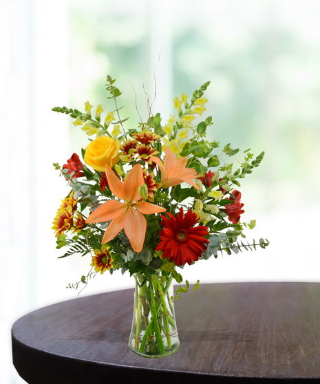 Bright fall flower arrangement with orange lilies, yellow roses, red gerberas, and seasonal greens in a clear vase