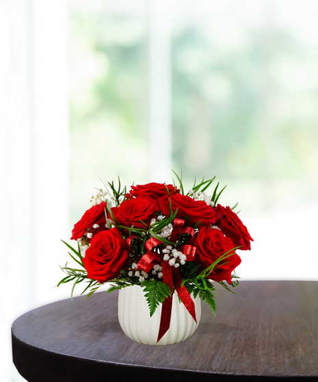 Put A Bow On It floral arrangement featuring lush red roses, white baby's breath, green foliage, and a sparkling red bow in a white ribbed vase