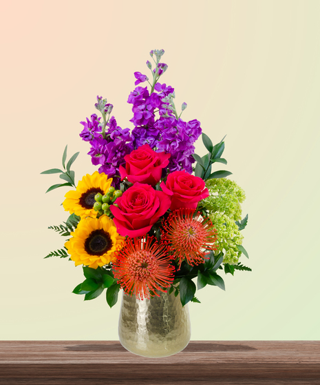 Dear Mom flower arrangement with hot pink roses, sunflowers, pincushion protea, purple stock, and green hydrangeas in a gold vase – perfect for Mother’s Day.