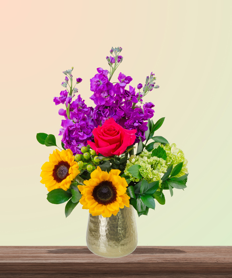 Dear Mom flower arrangement with hot pink roses, sunflowers, pincushion protea, purple stock, and green hydrangeas in a gold vase – perfect for Mother’s Day.