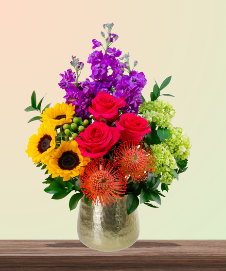 Dear Mom flower arrangement with hot pink roses, sunflowers, pincushion protea, purple stock, and green hydrangeas in a gold vase – perfect for Mother’s Day.