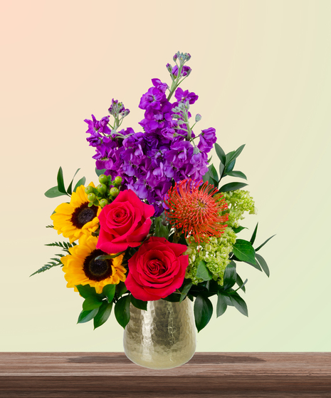 Dear Mom flower arrangement with hot pink roses, sunflowers, pincushion protea, purple stock, and green hydrangeas in a gold vase – perfect for Mother’s Day.