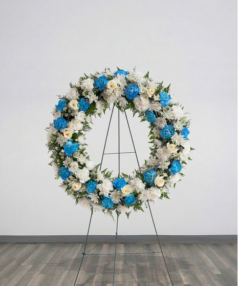 Sympathy wreath of white carnations, blue carnations, and ivory roses displayed on a metal easel – Touch of Color Collection by Billy Heroman’s
