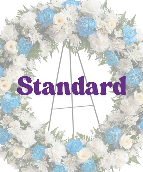 Sympathy wreath of white carnations, blue carnations, and ivory roses displayed on a metal easel – Touch of Color Collection by Billy Heroman’s