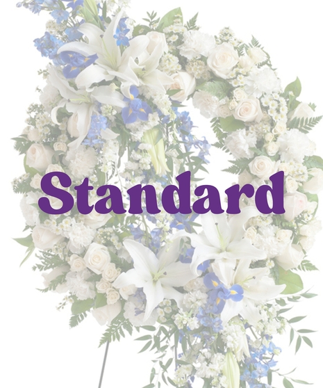 Elegant sympathy wreath with cascading white lilies, blue delphinium, ivory roses, and greenery on a metal easel – Touch of Color Collection by Billy Heroman’s