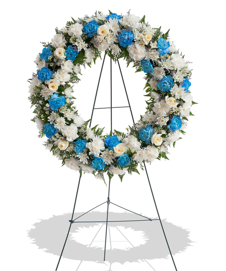 Sympathy wreath of white carnations, blue carnations, and ivory roses displayed on a metal easel – Touch of Color Collection by Billy Heroman’s