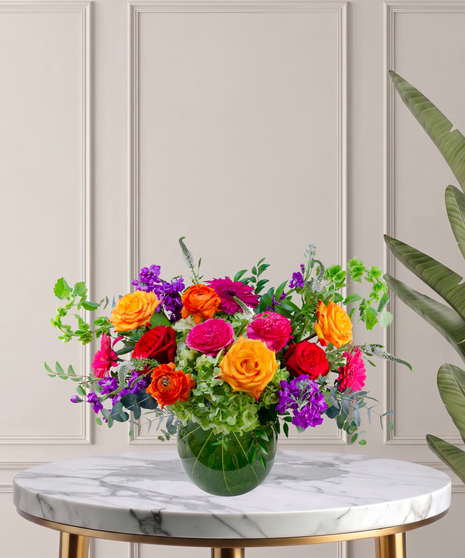 Zara floral arrangement featuring jewel-toned roses, ranunculus, hydrangeas, and gerbera daisies in a round glass vase with tropical leaf lining.