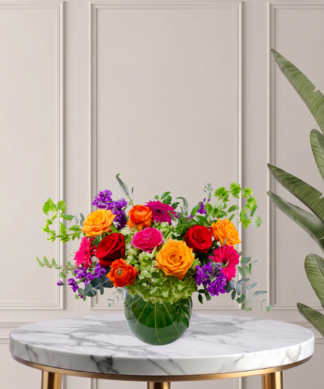 Zara floral arrangement featuring jewel-toned roses, ranunculus, hydrangeas, and gerbera daisies in a round glass vase with tropical leaf lining.