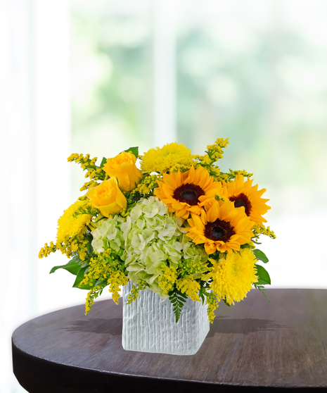 Lemon Drop floral arrangement with sunflowers, yellow roses, green hydrangeas, and solidago in a modern white vase on a wooden table.