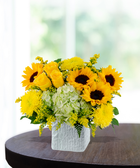 Lemon Drop floral arrangement with sunflowers, yellow roses, green hydrangeas, and solidago in a modern white vase on a wooden table.