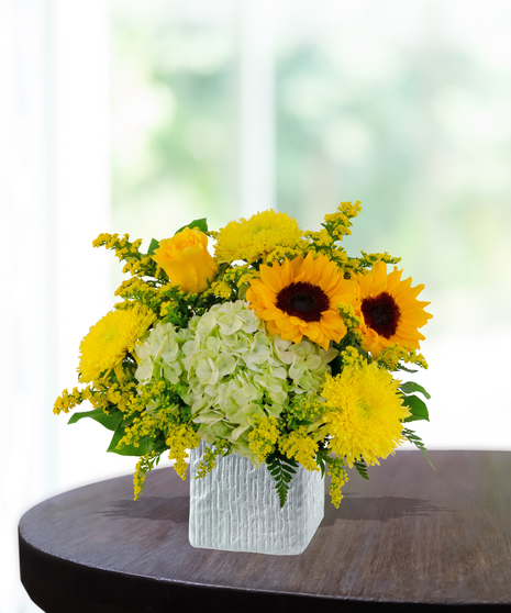 Lemon Drop floral arrangement with sunflowers, yellow roses, green hydrangeas, and solidago in a modern white vase on a wooden table.